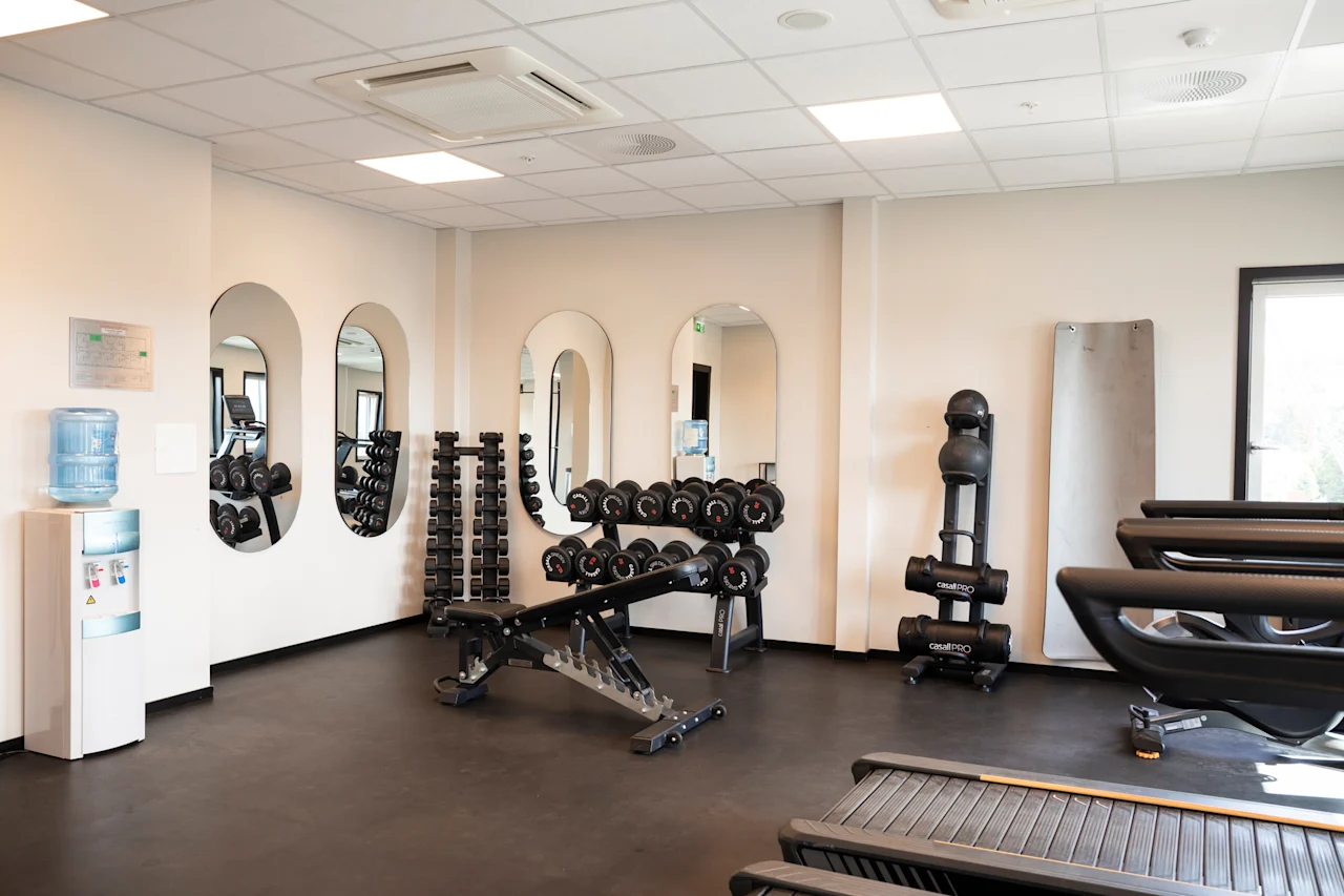 Et lyst og moderne treningsstudio med diverse treningsutstyr inkludert manualer, en treningsbenk og tredemøller. En vanndispenser er også synlig.
