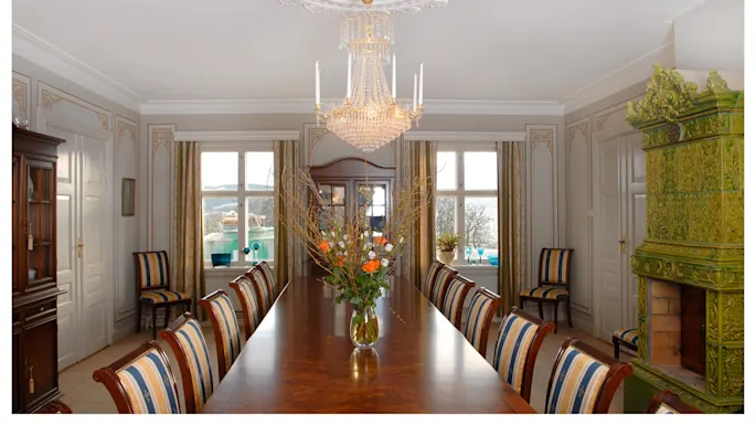 A long wooden table with a floral vase sits under a chandelier in an ornate dining room with striped chairs, decorative walls, large windows, and a green ceramic stove.