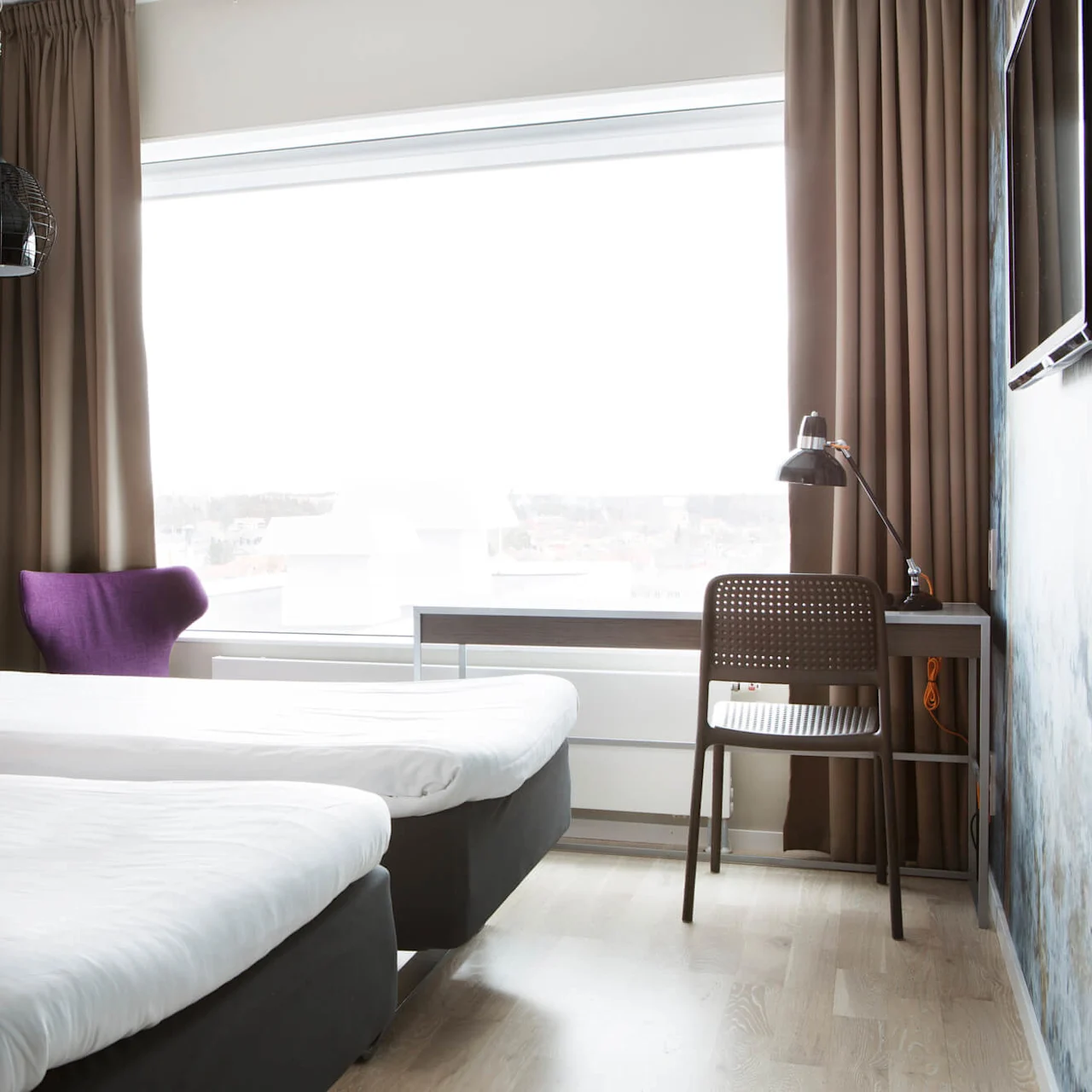 To pænt redte enkeltsenge står i et lyst hotelværelse med lyst trægulv. En lilla stol, skrivebord og lampe står nær et stort vindue med brune gardiner.