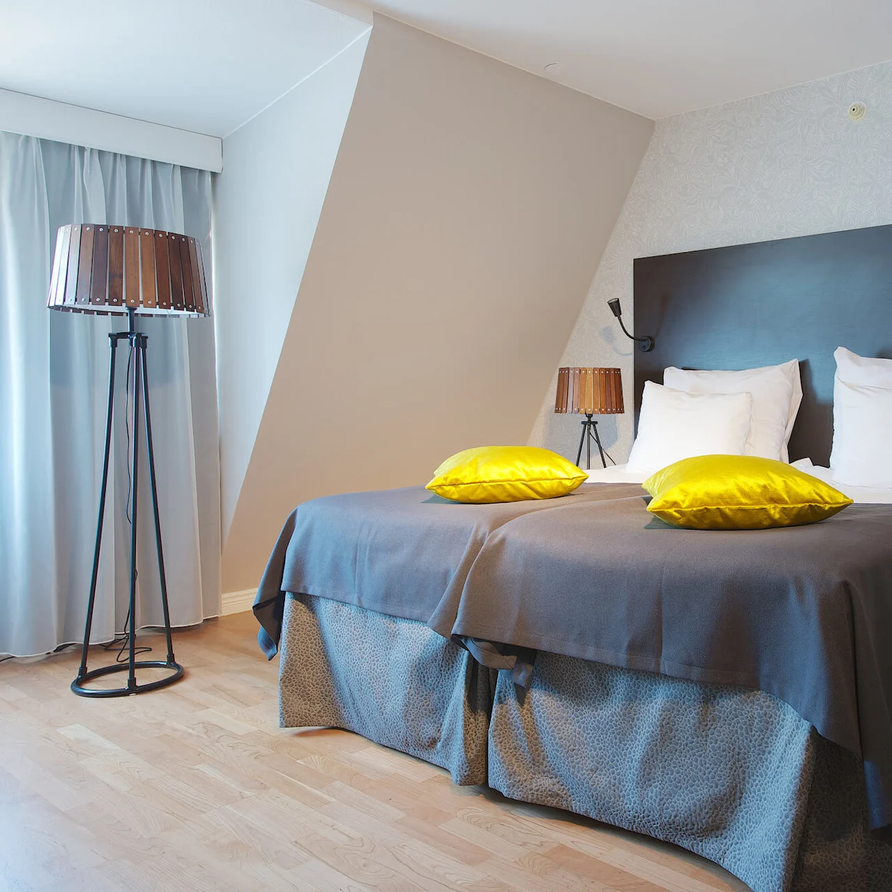 A neatly made bed with grey and yellow bedding in a bedroom, featuring a standing lamp and two tabletop lamps.