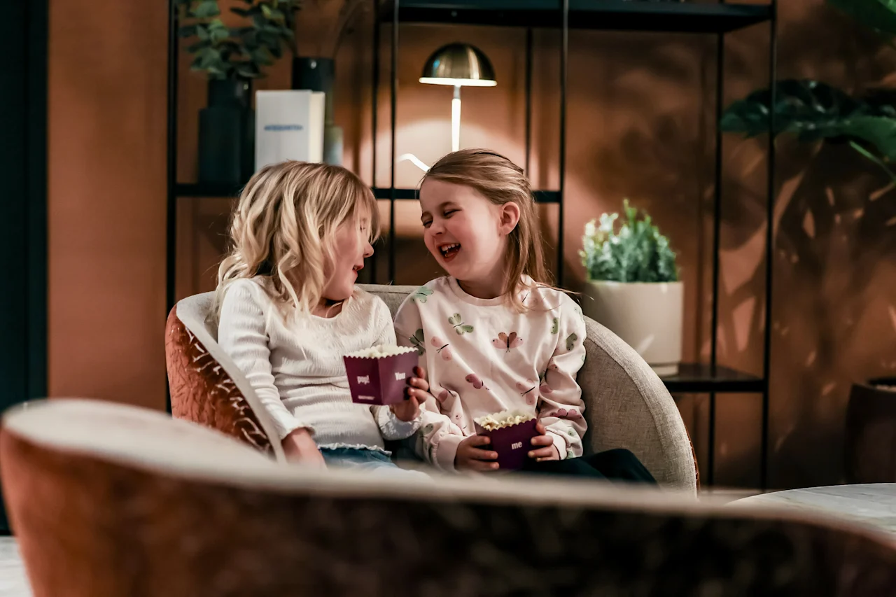 Two children sit close on a couch, laughing and holding small popcorn boxes. The room is warmly lit, with plants and a lamp in the background.