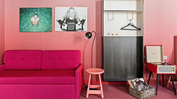 Rosa sofa står mot en mønstret vegg med fotografier over. I nærheten står en svart gulvlampe, rosa krakk og åpent garderobeskap med kleshengere. En Crosley-platespiller spiller ved siden av en kasse med vinylplater.
