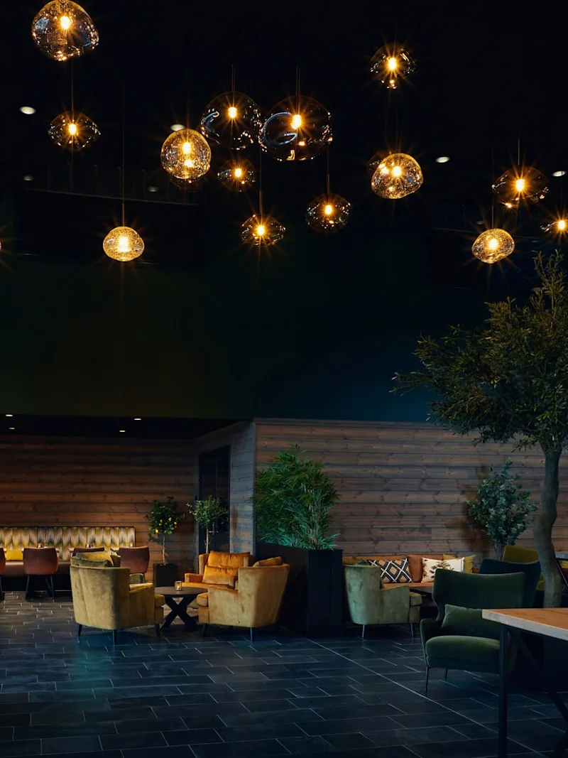 Hängande lampor lyser upp en mysig lounge med sammetsstolar, träbord och inomhusträd, vilket skapar en varm atmosfär. Rummet har träv väggar och kaklat golv.