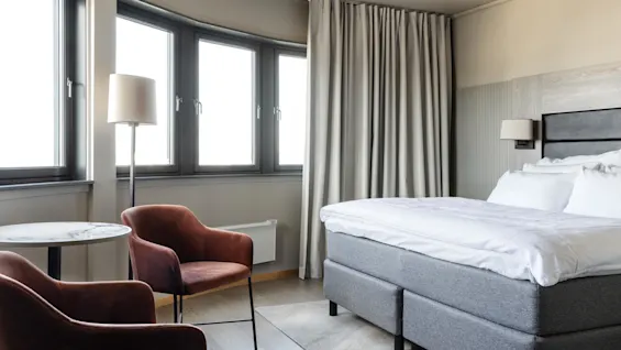 A neatly made double bed with white linens stands beside a small seating area with two orange chairs and a round table, near large windows with curtains.
