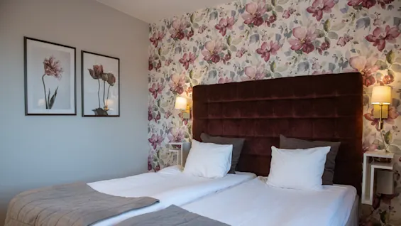 A neatly arranged bed sits against a floral wallpapered wall, with brown headboard and two framed floral artworks beside it, in a softly-lit bedroom setting.