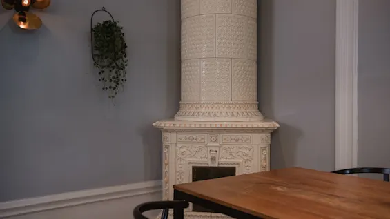 Elegant kakelugn står inaktiv bredvid ett träbord i ett mysigt rum; en liten vägglampa och hängande växt pryder de ljusgrå väggarna.