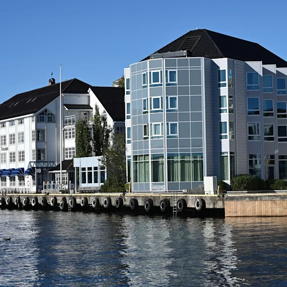 Et hotell med blå markiser og en moderne glassbygning ved vannkanten på en solrik dag. Teksten lyder Hotel.