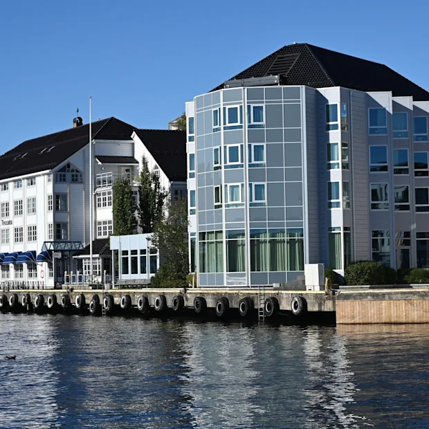 Et hotell med blå markiser og en moderne glassbygning ved vannkanten på en solrik dag. Teksten lyder Hotel.