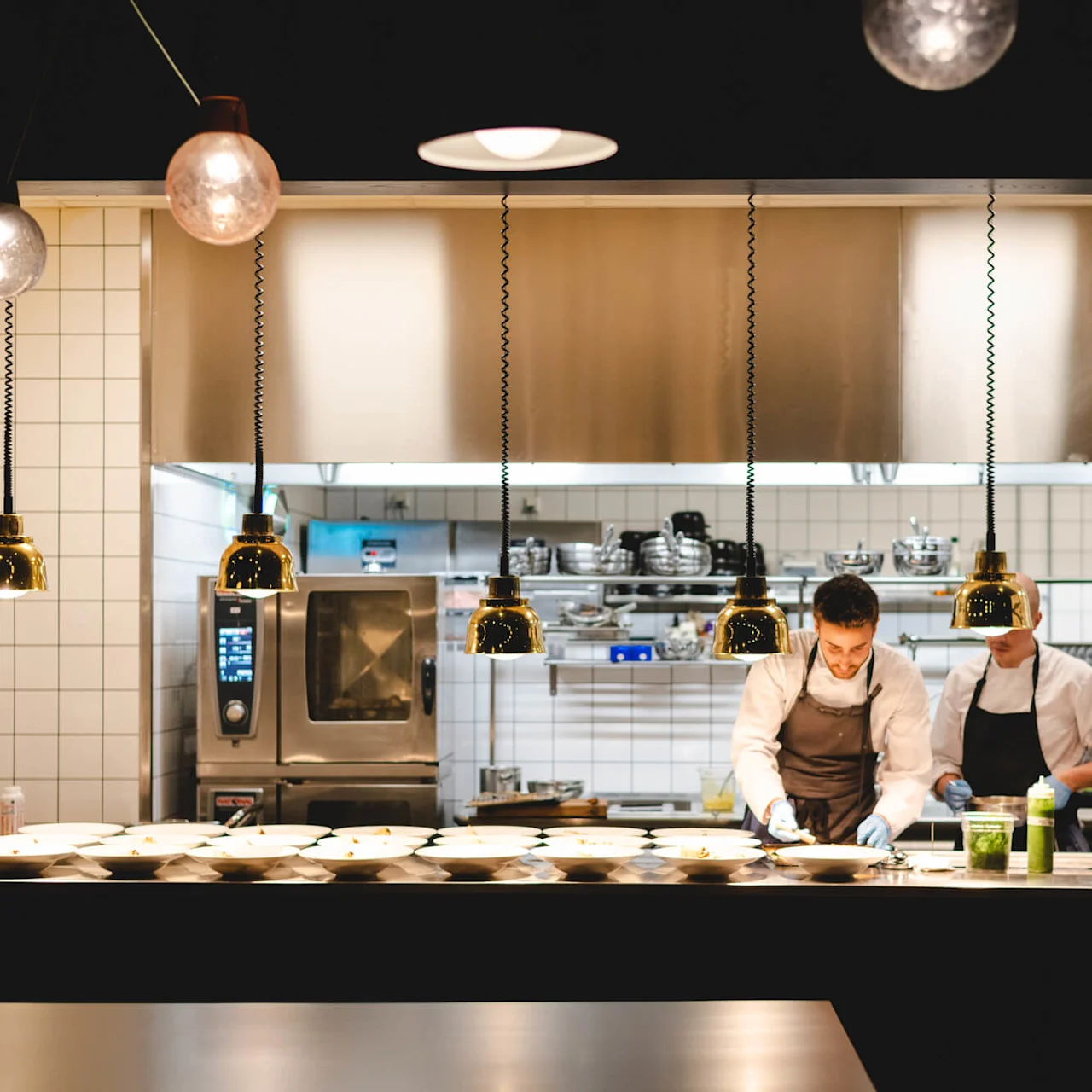 Kockar förbereder måltider i ett professionellt kök. Ljusa hängande lampor lyser upp arbetsområdet, och rostfria apparater och hyllor med köksutrustning skapar en hektisk, organiserad miljö.