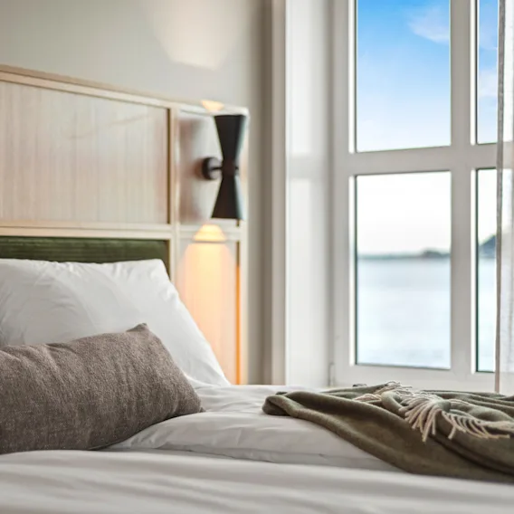 Et koselig hotellrom med en seng, puter og et teppe, med et stort vindu som vender ut mot et rolig vann under en blå himmel.