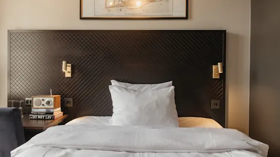 Ett mysigt hotellrum på Home Hotel Grand, Helsingborg, med en bekväm säng, sängbord med radio och böcker, samt en karta på väggen. MIT LIV