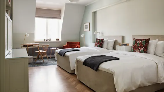 To pent oppredde senger med fargerike puter står i et mykt opplyst, moderne rom med et skrivebord, oransje lenestol og vindu, og skaper en koselig, innbydende atmosfære.