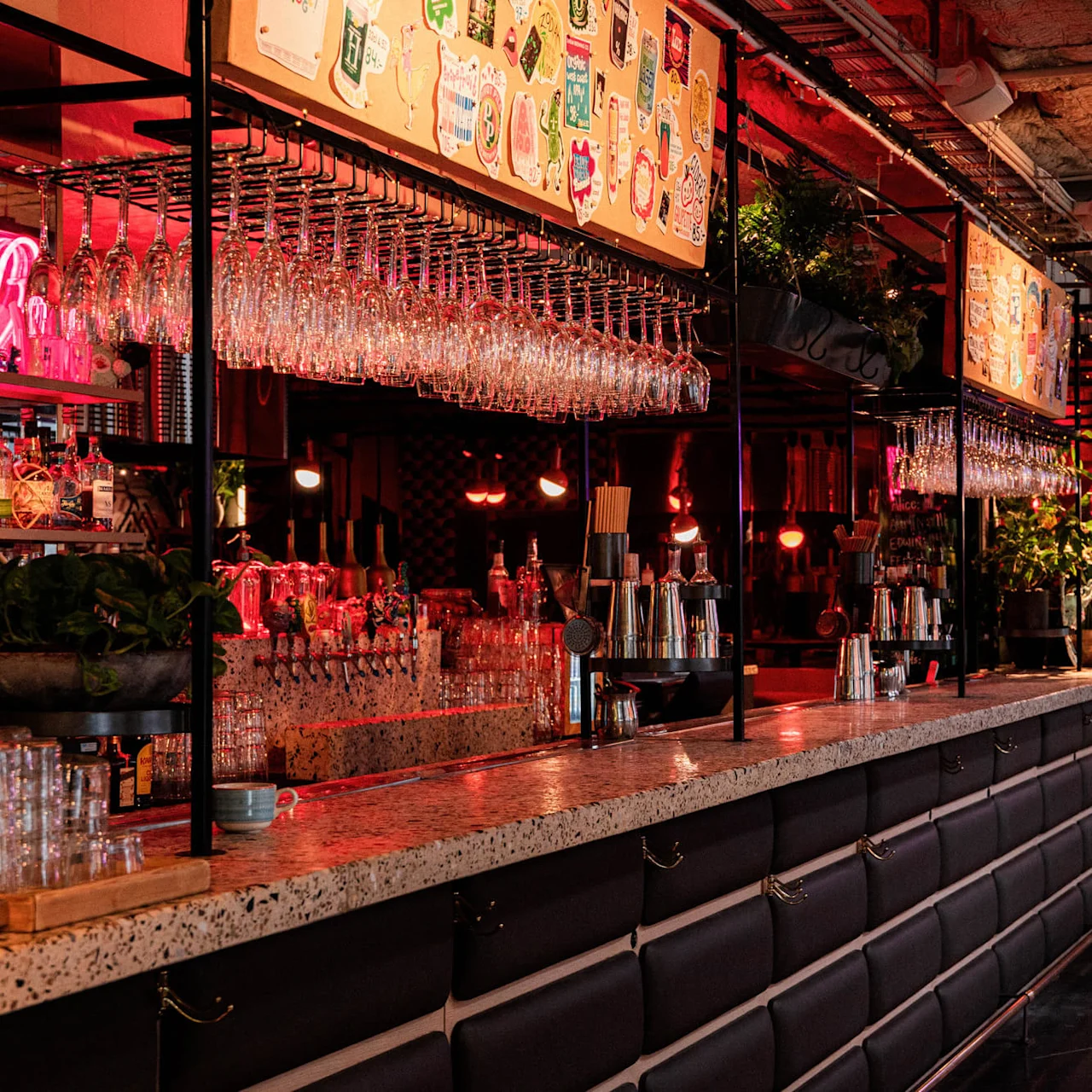 Bar counter lined with hanging glasses and cocktail shakers, situated in a dimly lit room with red neon lights. People sit at a table in the background.