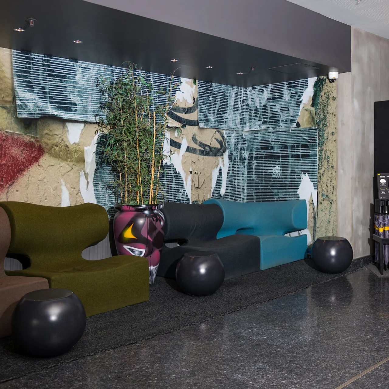 Colorful, modern lounge chairs sit against an abstract mural. A large potted plant occupies the center. To the right, a vending display reads "COMFORT DELI" offering snacks and drinks.