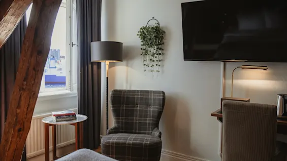 Et hyggeligt hotelværelse med en ternet lænestol, en gulvlampe og et tv. Et vindue giver udsigt over et skib på vandet.