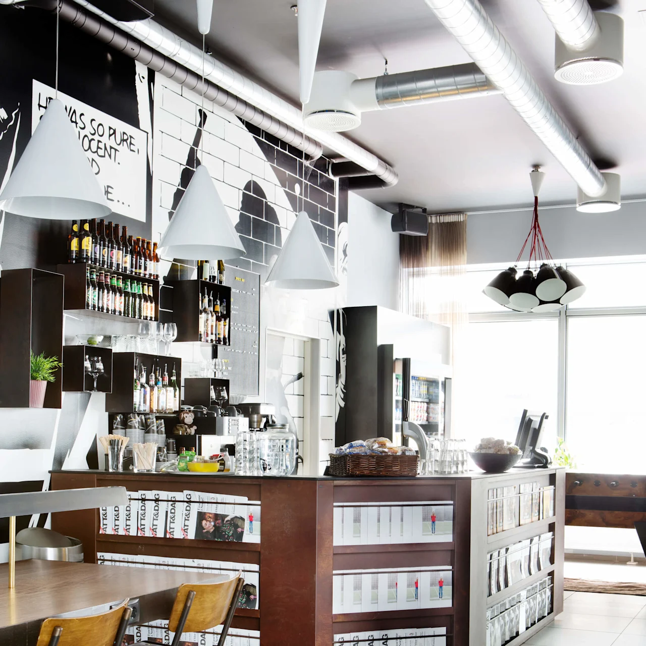 A modern cafe bar features shelves stocked with bottles behind the counter, with white cone-shaped lights hanging above. Magazines are displayed beneath. Wall text reads: "He was so pure, innocent." 