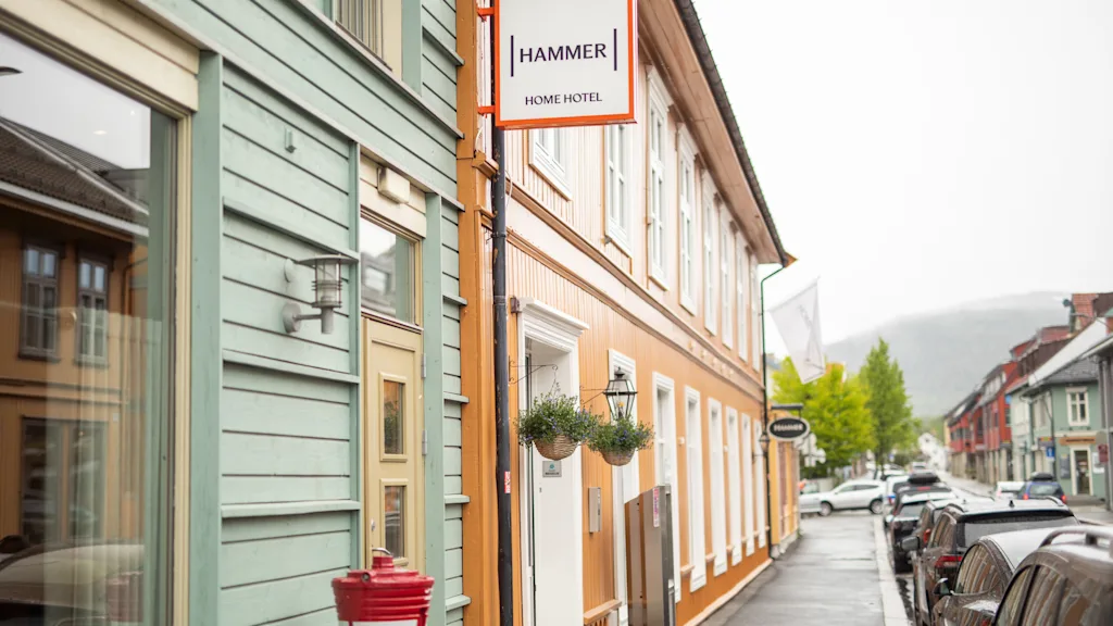 Bygning skilt med teksten "HAMMER HOME HOTEL" på en oransje bygning. Toetasjes bygninger langs en våt gate med parkerte biler. Trær synlige i bakgrunnen.
