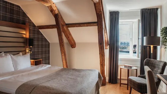 Et varmt og indbydende hotelværelse med en komfortabel seng, rustikke træbjælker og et vindue med udsigt.