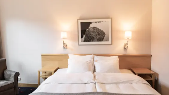 Et koselig hotellrom med en pent oppredd seng, sengegavl i tre, nattbordslamper og en lenestol, som inviterer til avslapning.