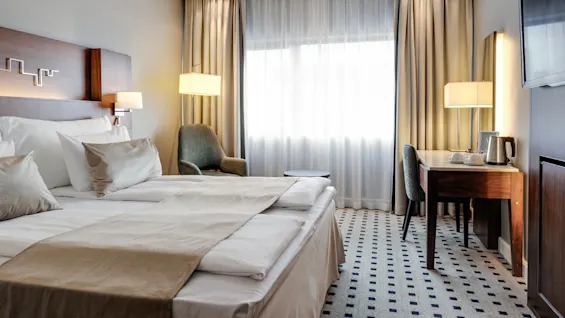 A neatly made double bed with beige linens sits in a well-lit hotel room. Nearby, a desk with a lamp, chair, and kettle completes the cozy, modern decor.