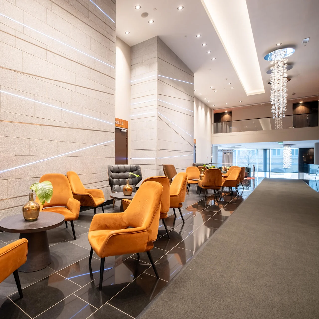 Orange chairs surround black tables in a modern lobby, featuring a tall stone wall with horizontal lines, a chandelier, and door labeled "HALL 1". 