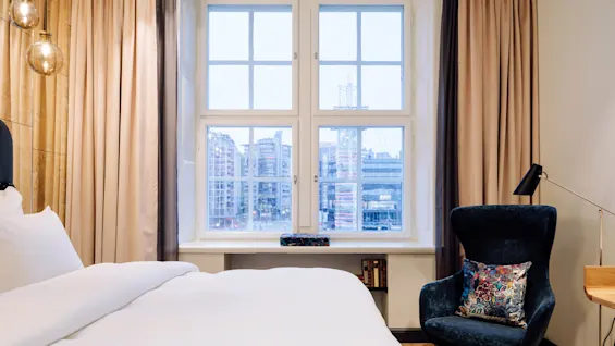 Bed covered in white sheets beside a large window with city view, surrounded by beige curtains; a dark blue armchair with a colorful pillow, and a desk lamp nearby.