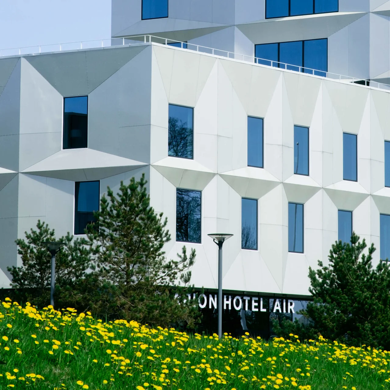 En moderne bygning med geometrisk hvit fasade og blå vinduer, beliggende ved siden av trær og gule blomster. Delvis synlig tekst: "ON HOTEL AIR".