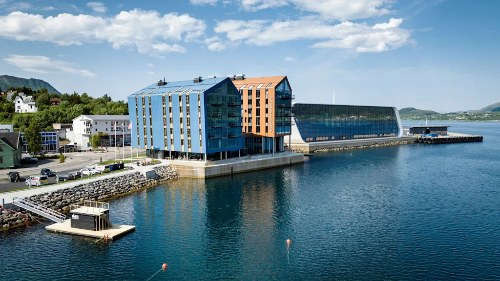 A modern, colorful building with a slanted roof sits by a calm waterfront, surrounded by clear skies and distant green hills. Nearby, a small floating dock features the text "NAUST".