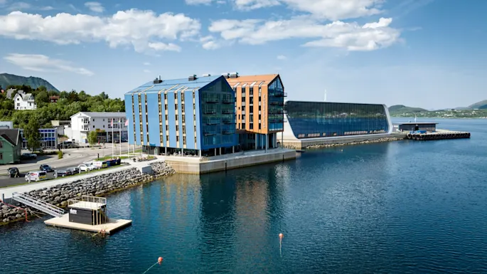 A modern, colorful building with a slanted roof sits by a calm waterfront, surrounded by clear skies and distant green hills. Nearby, a small floating dock features the text "NAUST".