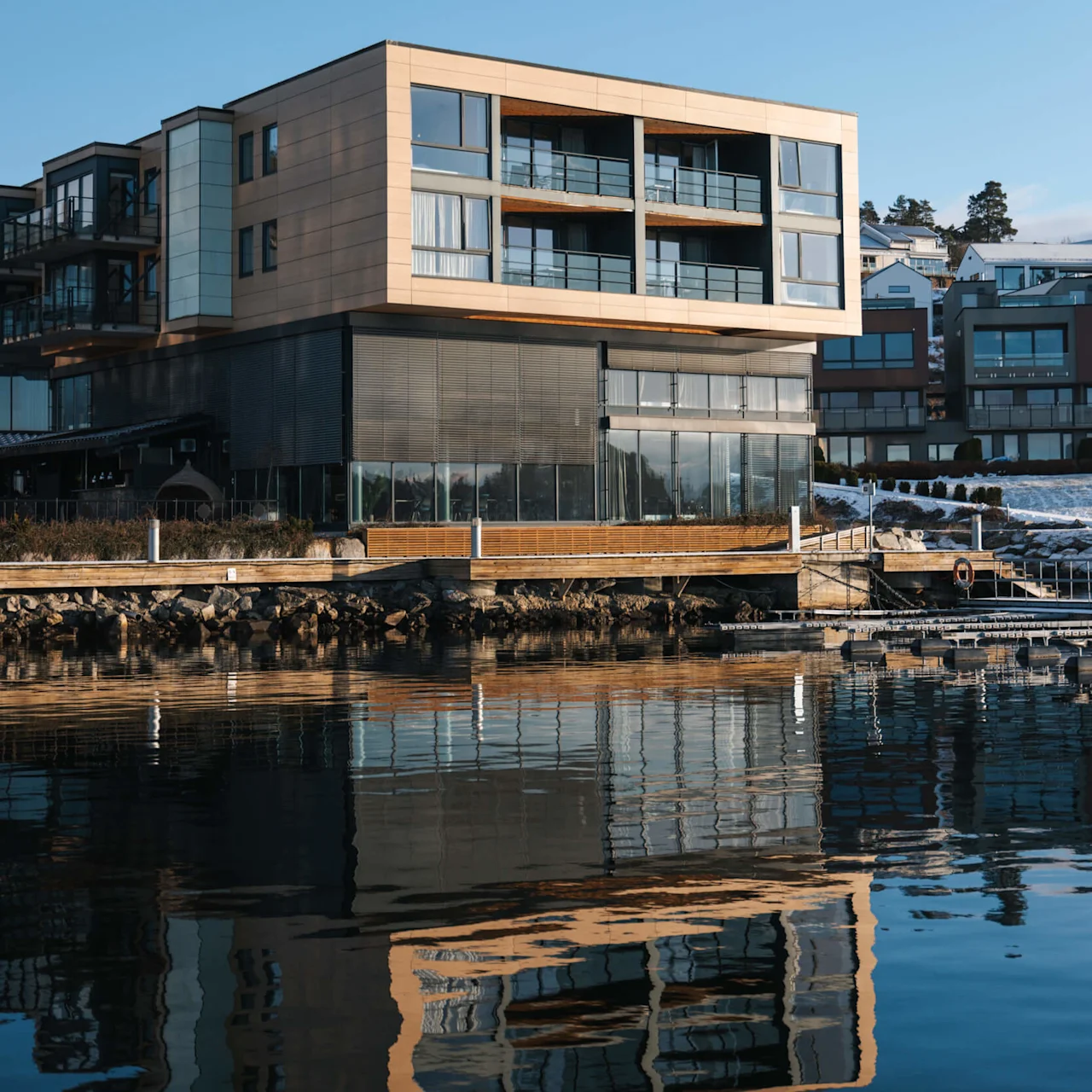 En moderne, fleretasje bygning reflekteres i rolig vann, omgitt av andre bygninger under en klar blå himmel.