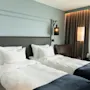 Et stilfullt hotellrom med to senger, skarpe hvite sengetøy, blå pledd og en lyseblå vegg. Myk belysning og en kaffemaskin.