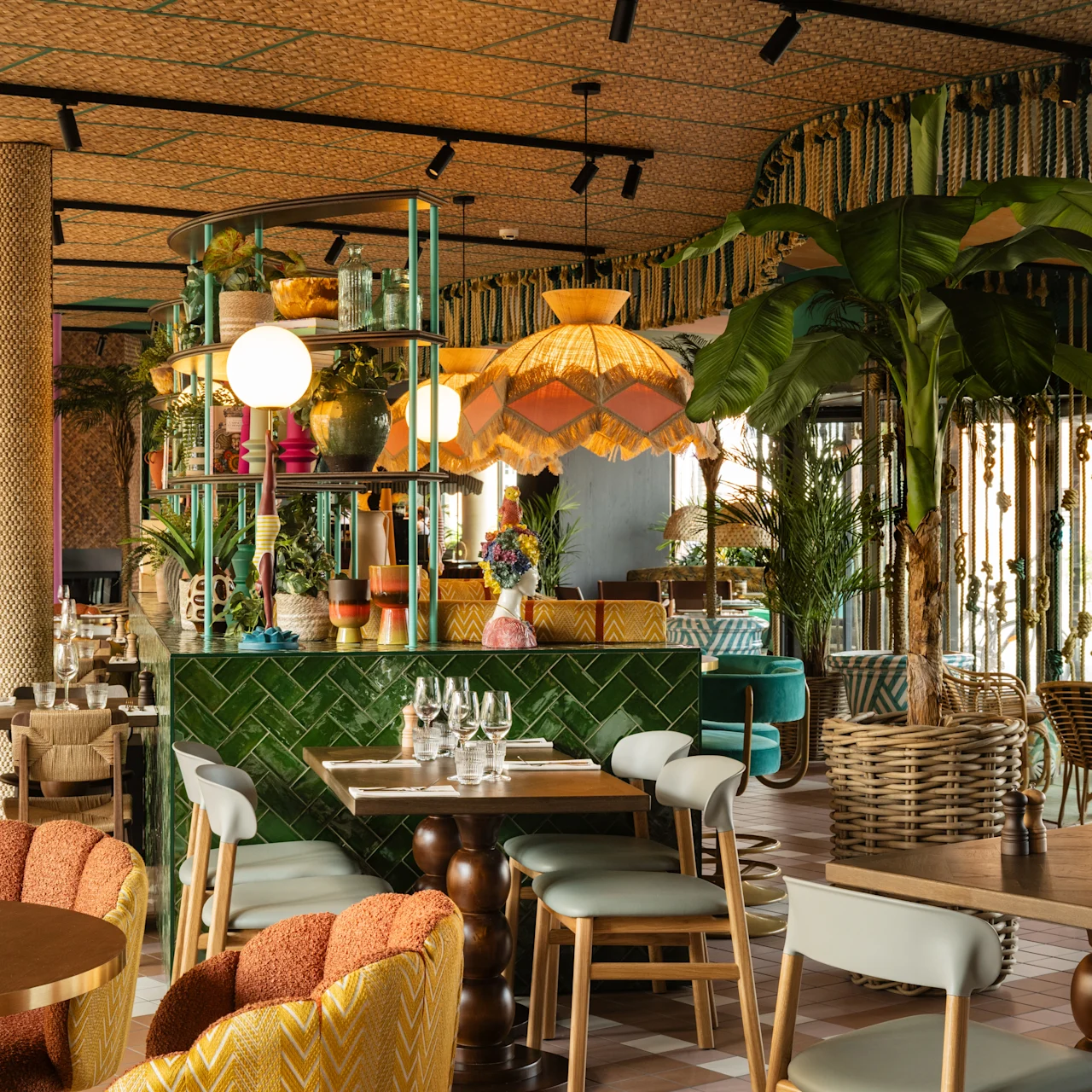 A restaurant interior features colorful, eclectic decor with lush plants, wooden furniture, and soft lighting. Tables are set with glasses and utensils, creating a welcoming dining atmosphere.