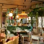 En restaurantindretning har farverig, eklektisk interiør med frodige planter, træmøbler og blød belysning. Bordene er sat med glas og bestik, hvilket skaber en indbydende spisestemning.