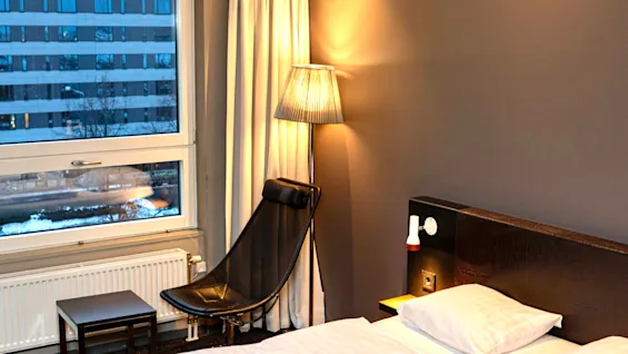 Ett mysigt hotellrum med en bekväm säng, en stilren stol och varm belysning, med utsikt över staden utanför.