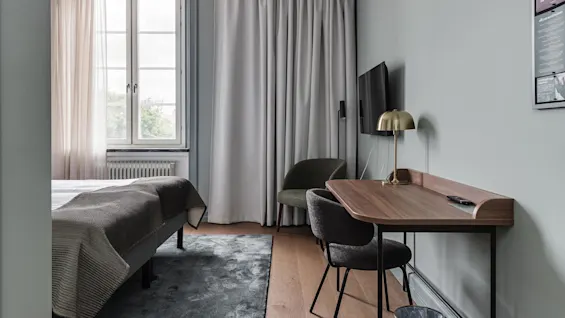 A modern room with a bed, TV, desk, and chair, draped windows, and subtle decor; serene and organised.
