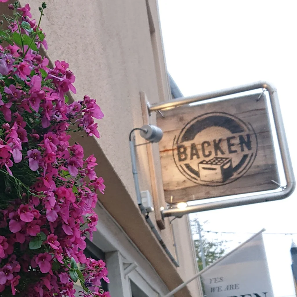 Lyserøde blomster falder fra en plantekasse ved siden af et træskilt og et flag, der siger  BACKEN