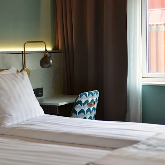 A neatly made bed with white linens is next to a window. A patterned chair faces a small desk with a lamp. Curtains partly cover the window.