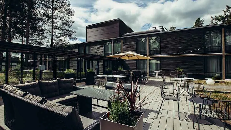 Patio furniture sits on a wooden deck, shaded by an umbrella, with a modern building backdrop.