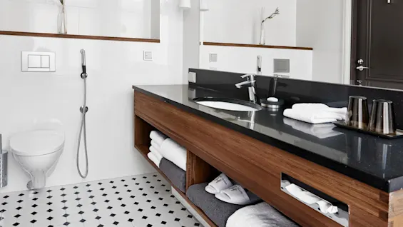 Badeværelse med sort bordplade, vask og vandhane. Skab i træ nedenunder rummer sammenfoldede håndklæder og hjemmesko. Væghængt toilet med bidetbruser. Minimalistisk indretning med glasvaser på en hylde.