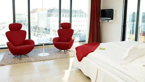Two red armchairs face a small white table on a rug, in a bright room with large windows overlooking a cityscape. A bed with red accents is nearby.