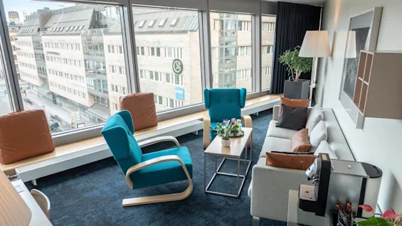 Ett modernt rum har en vit soffa och blå stolar runt ett litet bord med växter, med utsikt över en gata genom stora fönster, vilket avslöjar intilliggande byggnader.