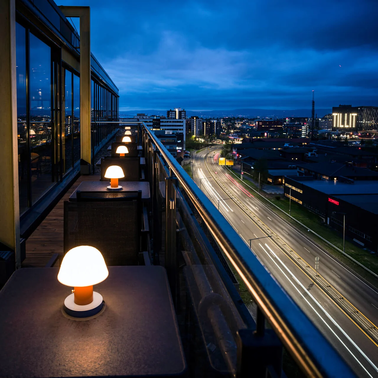 Outdoor terrace tables with glowing lamps line a high balcony overlooking a cityscape at night. In the distance, the word "TILIT" is illuminated on a building.