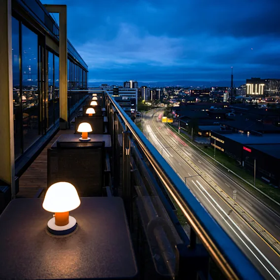 Utendørs terrassebord med glødende lamper står langs en høy balkong med utsikt over bylandskapet om natten. I det fjerne er ordet "TILIT" opplyst på en bygning.