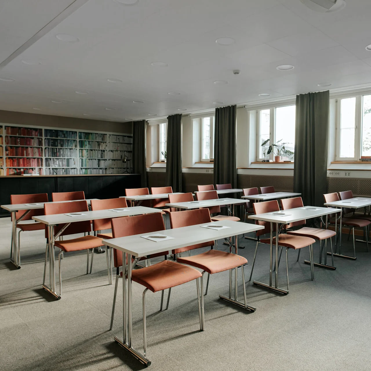 Empty classroom with rows of tables and chairs, bookshelf on one side, large windows with curtains.