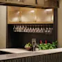 A glass of champagne sits on a modern bar counter. Hanging glasses and bottles are neatly arranged on shelves, surrounded by plants and fruit, creating an elegant atmosphere.
