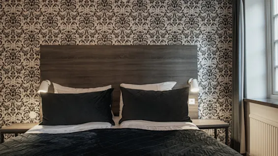 Ett bekvämt hotellrum med en mörk säng, mönstrad tapet och sänglampor, som erbjuder en avkopplande atmosfär.