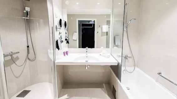 A modern bathroom with a clear glass shower partition, a sink, lit mirrors, and a bathtub, in a tiled, well-lit space.