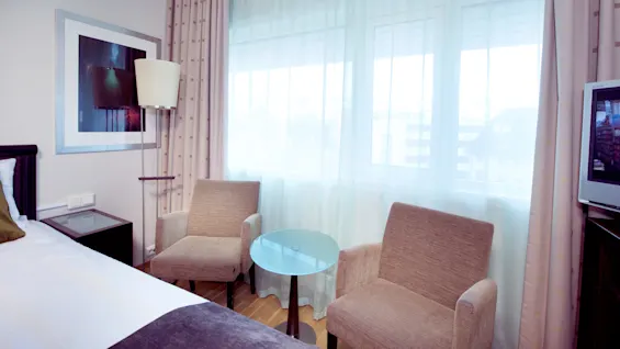 A hotel room with a bed, two armchairs, a small table, and a television, bright daylight streaming through sheer curtains.