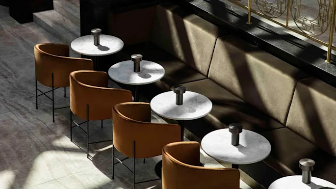 Tables with round marble tops are placed in a row, accompanied by brown upholstered chairs. The setting is a stylish lounge area with soft lighting.