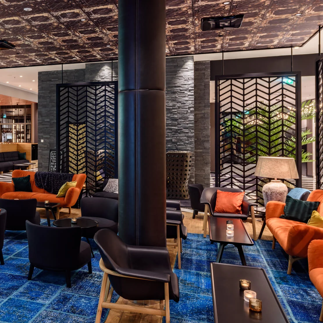 A stylish bar features leather stools and a well-stocked backlit counter. Orange and black seating with vibrant cushions sit on a blue-patterned carpet, creating a modern, cozy atmosphere.